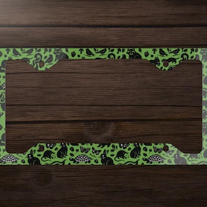 May include: Green license plate frame with a repeating pattern of black animal silhouettes, including cats, rabbits, and turtles. The frame has a rectangular shape with rounded corners and is set against a dark wood background.