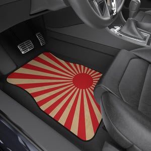 Puede incluir: Alfombrilla para coche con diseño de rayos de sol rojos sobre fondo beige. La alfombrilla, con borde negro, está colocada en el espacio para los pies del conductor. El interior del coche es negro, con pedales visibles.