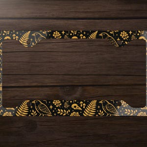 Puede incluir: Un marco de matrícula con fondo negro y un diseño dorado con cuervos, helechos, setas y elementos florales. El marco tiene un borde transparente y está diseñado para una placa de matrícula estándar.