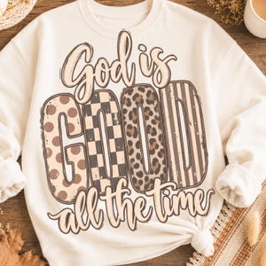 Puede incluir: Una sudadera de cuello redondo color crema con el texto "God is GOOD all the time" en una fuente estilizada. La palabra "GOOD" está rellena con cuatro patrones distintos: lunares marrones, un tablero de ajedrez blanco y negro, estampado de leopardo y rayas verticales.