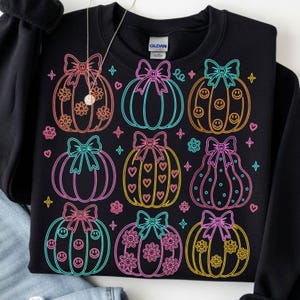 May include: A black crewneck sweatshirt featuring a neon-style graphic print of nine pumpkins arranged in a grid. Each pumpkin is outlined in bright colors and decorated with bows, smiley faces, hearts, or flowers, accented by small stars and hearts.