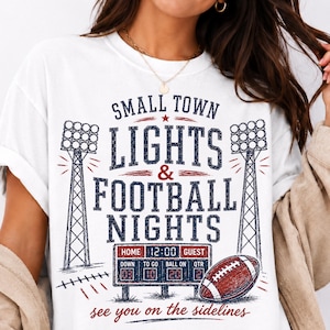 May include: A white t-shirt featuring a graphic print with the text "SMALL TOWN LIGHTS & FOOTBALL NIGHTS" and "see you on the sidelines". The design includes two stadium light towers, a scoreboard showing game stats, and a brown football.