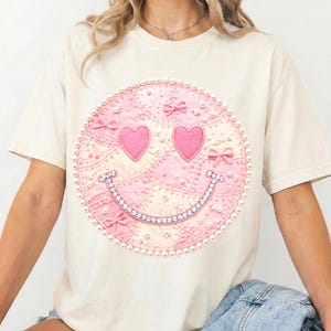 以下が含まれることがあります： クリーム色のTシャツ。大きなピンク色のスマイリーフェイスのデザイン。スマイリーフェイスはハート型の目、宝石をちりばめた笑顔で、パールとリボンで囲まれています。デザインはパッチワークスタイルです。