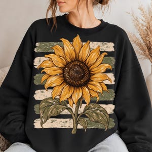 Puede incluir: Una sudadera negra de cuello redondo con un gráfico grande y centrado de un girasol amarillo con el centro marrón oscuro y hojas verdes. El girasol está superpuesto sobre un fondo de rayas horizontales desgastadas en color blanco roto y verde oliva.