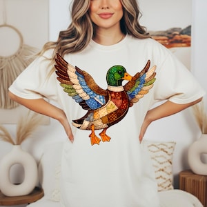 以下が含まれることがあります： クリーム色のTシャツ。カラフルなアヒルのグラフィックが描かれており、翼を広げています。アヒルのデザインは、茶色、青、緑、オレンジの色合いのパッチワークパターンを取り入れています。カジュアルな普段着です。
