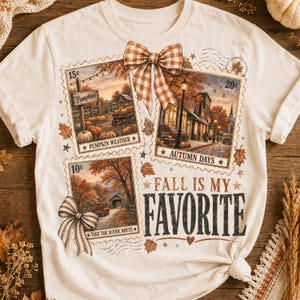 May include: A cream-colored t-shirt featuring three rectangular postage stamp graphics with autumn scenes. The stamps show a pumpkin patch, a town street, and a country road. A plaid bow sits at the top, and the text reads "FALL IS MY FAVORITE."