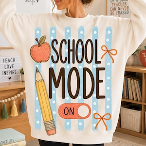 Peut inclure: Un sweat-shirt à col rond couleur crème avec un imprimé graphique à rayures verticales bleu clair à pois. Le motif comprend une pomme rouge, un crayon jaune, des nœuds orange et le texte "SCHOOL MODE ON" en grandes lettres marron.