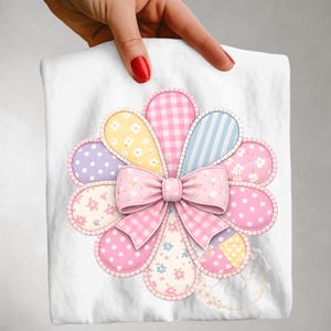 Könnte beinhalten: Ein weißes quadratisches Tuch mit einem großen, farbenfrohen Blumenmuster. Die Blume hat blütenblattförmige Abschnitte in Rosa, Gelb, Lila und Blau, mit Punkten, Vichy-Karo und floralen Mustern. In der Mitte befindet sich eine rosa Schleife.