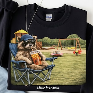 May include: Black sweatshirt featuring a raccoon wearing sunglasses and a baseball cap, relaxing in a blue folding chair with a drink. The graphic includes a playground scene and the text "i love here now".
