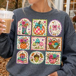Könnte beinhalten: Ein dunkelgraues Sweatshirt mit Rundhalsausschnitt und einem Raster aus neun farbenfrohen Illustrationen, darunter ein blumiger Cowboystiefel, ein karierter Kürbis mit blauer Schleife, eine Gans mit rosa Schleife, ein Blumenkrug, Pilze, ein Erntekorb, Bücher, eine Eichel und eine blumige Teetasse.