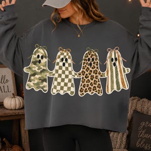 May include: A charcoal gray long-sleeve sweatshirt featuring a graphic of four ghosts holding hands. Each ghost has a different pattern: green camouflage, green and white checkerboard, brown leopard print, and green and white vertical stripes.