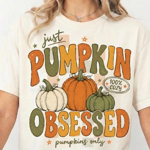 May include: A cream-colored t-shirt featuring a graphic print with the text "just PUMPKIN OBSESSED" and "pumpkins only". The design includes three pumpkins in white, orange, and green, a checkered background, and a "100% cozy" oval emblem.