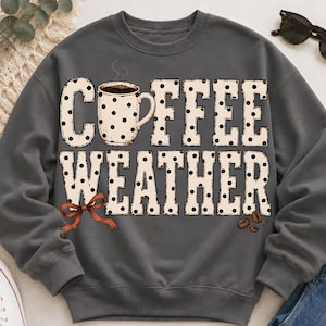 Könnte beinhalten: Ein anthrazitfarbenes Sweatshirt mit Rundhalsausschnitt und einem Grafikdruck mit dem Text COFFEE WEATHER in großen, cremefarbenen Buchstaben mit schwarzen Punkten. Eine cremefarbene Kaffeetasse, eine kleine orangefarbene Schleife und drei Kaffeebohnen.