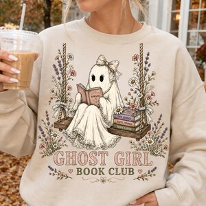 May include: A beige crewneck sweatshirt featuring a graphic of a ghost wearing a bow, sitting on a rope swing and reading a book. The design includes floral accents, a stack of books, and the text GHOST GIRL BOOK CLUB in pink and green.