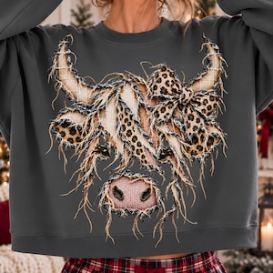 Puede incluir: Una sudadera de manga larga gris oscuro con un gran gr&aacute;fico de una vaca Highland. El dise&ntilde;o de la vaca est&aacute; compuesto por texturas deshilachadas tipo arpillera y patrones de leopardo, incluyendo un lazo de leopardo en la cabeza y nariz rosa.