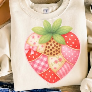 Puede incluir: Sudadera color crema con un diseño grande y colorido de fresa. La fresa está compuesta por varios patrones de patchwork, incluyendo lunares, cuadros y estampados florales, con un lazo rosa y hojas verdes.