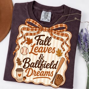 May include: A dark brown t-shirt featuring a graphic print of a plaid ribbon bow above a scalloped sign with the text Fall Leaves & Ballfield Dreams. The design includes illustrations of maple leaves, a baseball, a bat, a glove, and a coffee cup.