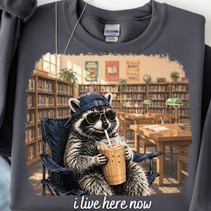 Puede incluir: Sudadera gris oscuro con un mapache con gafas de sol y una gorra de béisbol, sosteniendo un café helado. El mapache está sentado en una silla en una biblioteca. El texto "i live here now" está en la parte inferior.