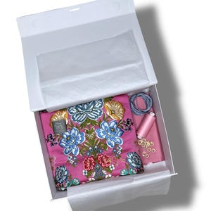May include: A white box containing a pink floral fabric, thread spools, and other sewing supplies. The fabric features a detailed floral pattern in blue, gold, and red. A small label is attached to the fabric. The box is lined with white tissue paper.