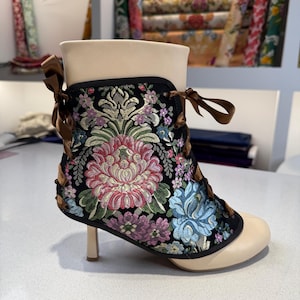 May include: Ankle boot with a cream-colored toe and heel, and a black upper adorned with a floral embroidery in pink, blue, and purple. The boot features a small heel and brown ribbon laces.