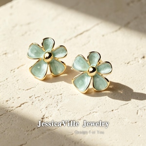 Op de afbeelding: Een paar bloemvormige oorstekers. Elke oorbel heeft een lichtgroene emaille bloem met goudkleurige rand en een goudkleurig midden. De oorbellen worden gepresenteerd op een getextureerd, lichtgekleurd oppervlak. De tekst "JessicaVille Jewelry" is zichtbaar.