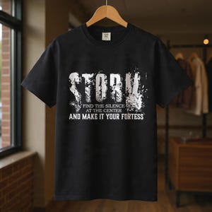 Pode incluir: Uma t-shirt preta de manga curta com uma impressão gráfica branca que apresenta a palavra STORM numa fonte desgastada e envelhecida. Abaixo, o texto diz: FIND THE SILENCE AT THE CENTER AND MAKE IT YOUR FORTRESS. Pendurada num cabide de madeira.