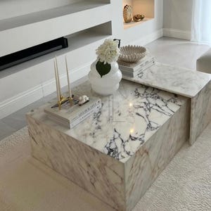 May include: A rectangular marble coffee table with a white and gray veined pattern. The table has a smaller, sliding top section. Decorative items include a white vase with flowers, candles, and books.