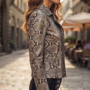 May include: A long-sleeved jacket featuring a grey and brown snakeskin pattern with a collared neckline and a zippered front closure. The jacket is paired with black pants, and the overall design includes visible side pockets with silver-toned zippers.