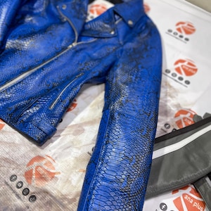 Puede incluir: Una chaqueta de motorista azul vibrante con un patrón texturizado de piel de serpiente en negro y azul. La chaqueta incluye cuello con muescas, cierre de cremallera frontal plateado, mangas largas y un cinturón con hebilla, con acabado en escamas.