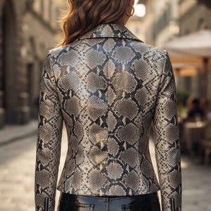 May include: A fitted jacket featuring a grey and black snakeskin pattern with a pointed collar, long sleeves, and vertical zippered cuffs. The jacket is worn over black, high-gloss, patent-finish trousers with visible back pockets and stitching.