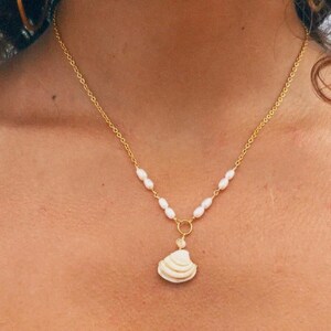 May include: A gold chain necklace featuring a series of small, white, oval-shaped freshwater pearls integrated into the links. The necklace includes a central pendant shaped like a white seashell, suspended from a small gold circular ring connector.