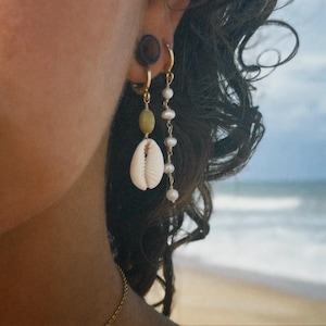 May include: Two gold-toned earrings. One earring features a small green bead and a white cowrie shell. The second earring is a long, delicate gold chain adorned with a series of small, round, white pearls hanging in a vertical line.