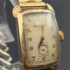 May include: A vintage Bulova wristwatch with a gold-toned rectangular case and a cream-coloured face. The watch features gold hands, a subdial, and gold-tone hour markers. The watch band is gold and silver.