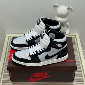 Puede incluir: Un par de zapatillas altas en blanco y negro. Las zapatillas presentan un diseño texturizado en la parte superior, cordones blancos y un logotipo de Nike negro. Se exhiben en una caja marrón con el logotipo de Nike.