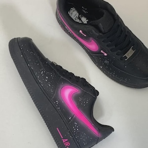 May include: Black sneakers with a pink Nike swoosh and accents. The shoes have white speckles and the word "AIR" in pink on the side. The sneakers are on a white surface.