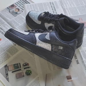 May include: Black sneakers with silver glitter accents. The shoes have a black sole, laces, and Nike swoosh. The sneakers are displayed on a newspaper background.