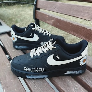 Pode incluir: Tênis pretos com detalhes brancos, incluindo um grande logotipo da Nike e cadarços. Os tênis apresentam um design de margarida e a palavra "POWERFUL" na lateral. As solas têm a inscrição "AIR". Os tênis são exibidos em um banco de madeira.