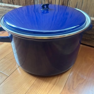 May include: A deep blue enamel cooking pot with a matching lid. The pot has two side handles and a silver trim. The lid features a small black handle. The pot is on a wooden surface.