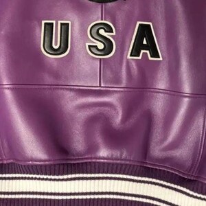 May include: A close-up of a purple leather jacket with the word "USA" in black and white lettering across the chest. The jacket has a ribbed hem with white and black stripes.