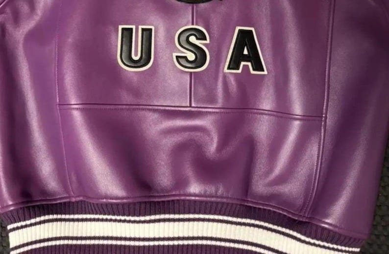 May include: A close-up of a purple leather jacket with the word "USA" in black and white lettering across the chest. The jacket has a ribbed hem with white and black stripes.