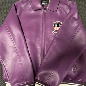 May include: A purple leather jacket with a zip-up front and a collared neckline. The jacket features white and purple striped cuffs and a patch on the chest with the text "AVIREX" and an American flag design.