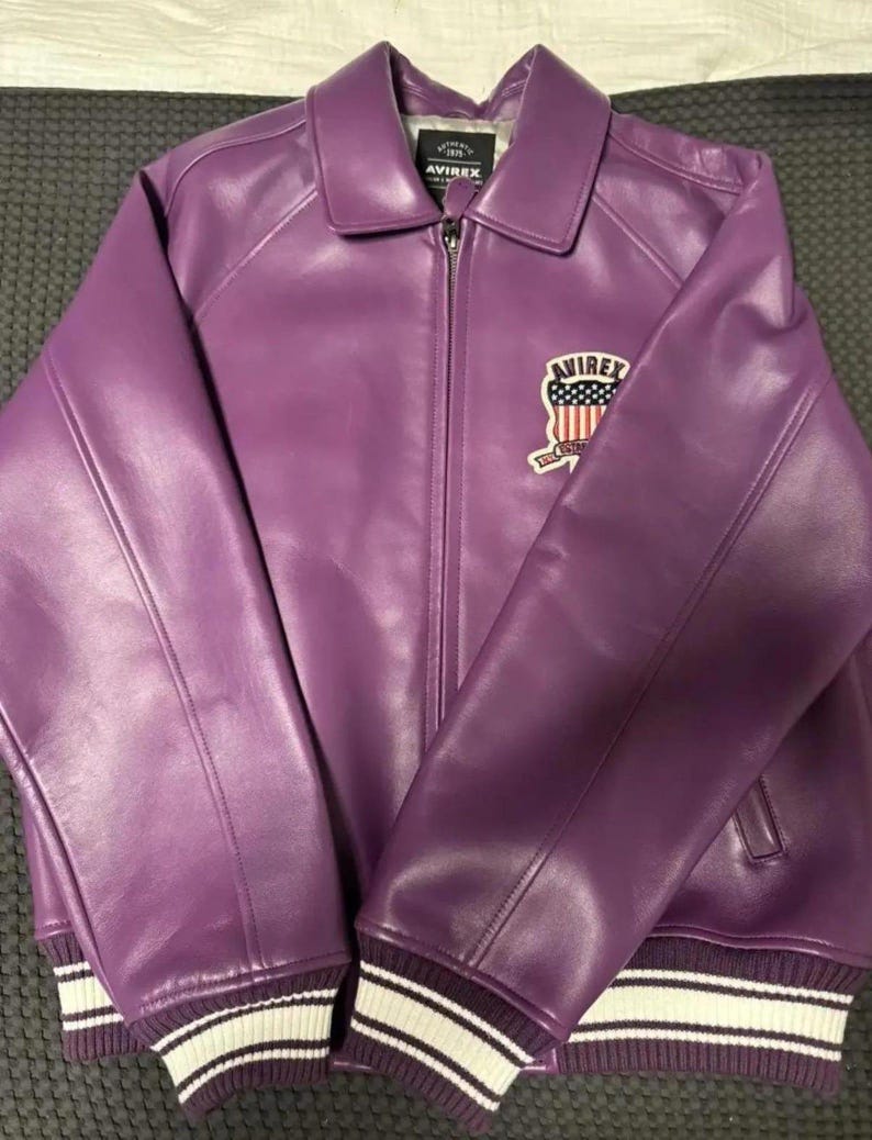 May include: A purple leather jacket with a zip-up front and a collared neckline. The jacket features white and purple striped cuffs and a patch on the chest with the text "AVIREX" and an American flag design.