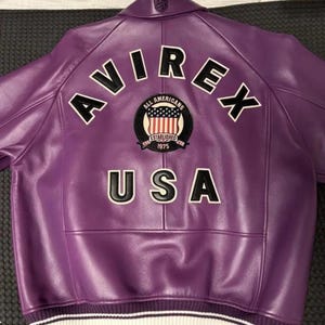 May include: A purple leather jacket with the word "AVIREX" arched across the upper back in white block letters. Below the word "AVIREX" is a patch with the words "ALL AMERICANS ESTABLISHED 1975 USA". The jacket has a white and black striped band at the waist.
