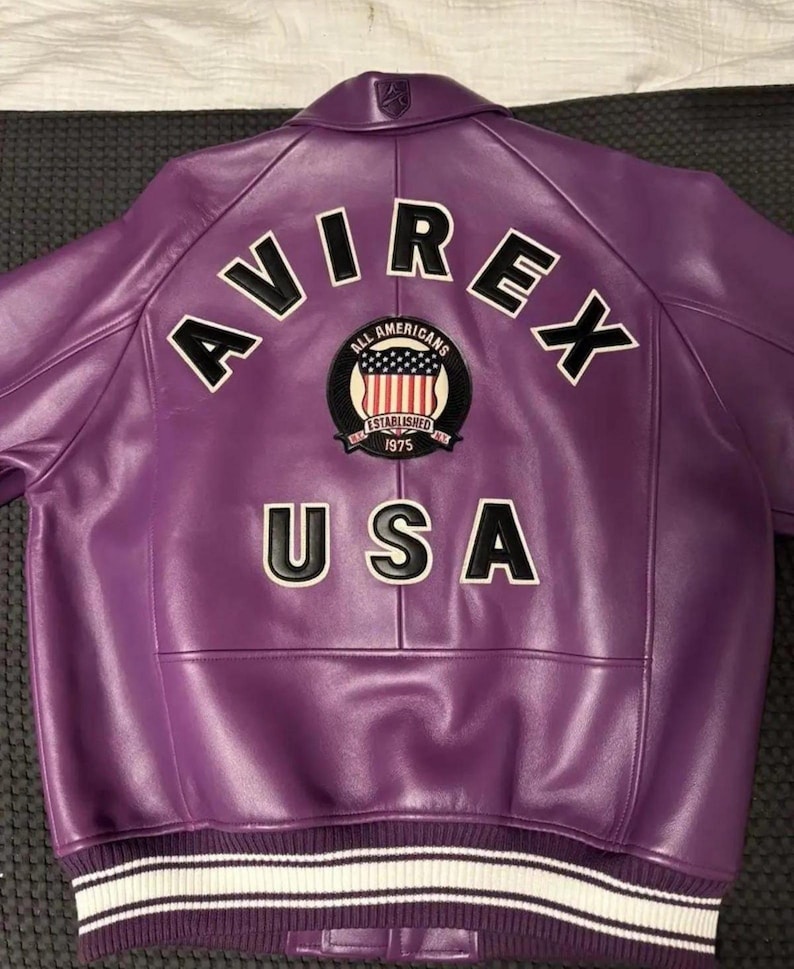 May include: A purple leather jacket with the word "AVIREX" arched across the upper back in white block letters. Below the word "AVIREX" is a patch with the words "ALL AMERICANS ESTABLISHED 1975 USA". The jacket has a white and black striped band at the waist.