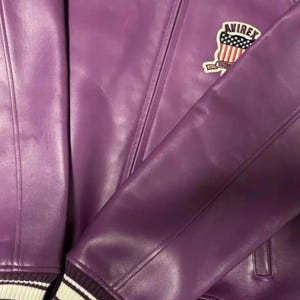 May include: A purple leather jacket with a classic design. The jacket features a patch with the word "AVIREX" and a design of the American flag. The sleeve cuff has white and black stripes. The jacket is made of a smooth, shiny material.
