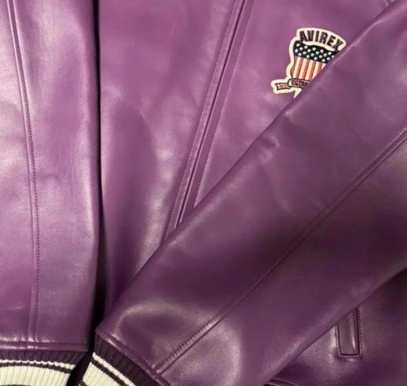 May include: A purple leather jacket with a classic design. The jacket features a patch with the word "AVIREX" and a design of the American flag. The sleeve cuff has white and black stripes. The jacket is made of a smooth, shiny material.