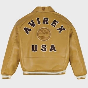 Puede incluir: Chaqueta de cuero color mostaza con la palabra "AVIREX" arqueada en la espalda en letras marrones, junto con un logotipo de &aacute;rbol y "USA". La chaqueta tiene cuello, pu&ntilde;os acanalados y dobladillo acanalado con rayas blancas.