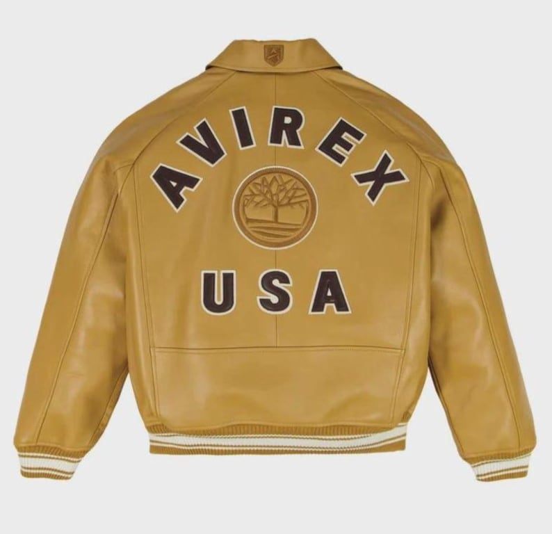 Puede incluir: Chaqueta de cuero color mostaza con la palabra "AVIREX" arqueada en la espalda en letras marrones, junto con un logotipo de &aacute;rbol y "USA". La chaqueta tiene cuello, pu&ntilde;os acanalados y dobladillo acanalado con rayas blancas.