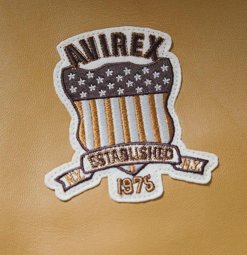Puede incluir: Parche bordado con la palabra "AVIREX" en la parte superior, sobre un dise&ntilde;o de escudo con estrellas y rayas. El parche incluye el texto "ESTABLISHED NY 1975" en una pancarta. El parche es blanco con detalles en marr&oacute;n, dorado y beige.