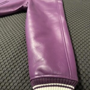 May include: A close-up of a purple leather jacket sleeve. The sleeve has a ribbed cuff with white stripes. A small patch is visible on the upper part of the jacket. The jacket is made of a smooth, shiny material.